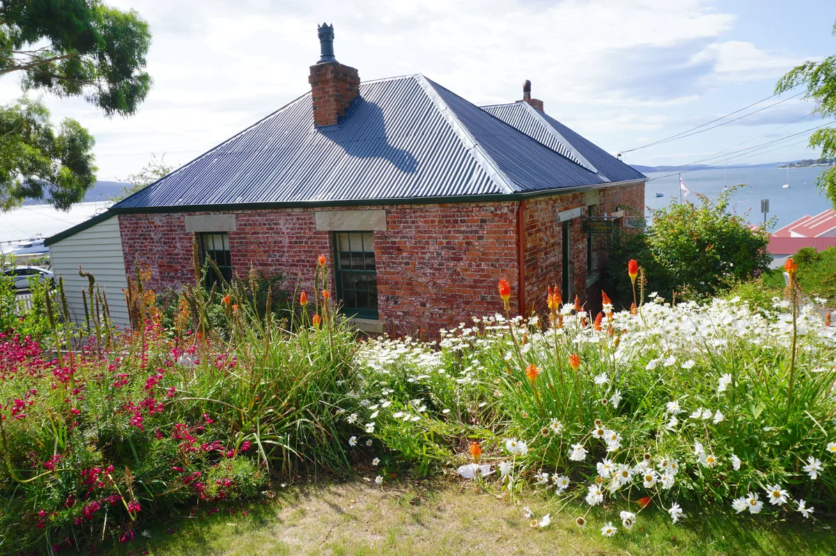 A brick cottage with sandstone lintels in Battery Point, Hobart, with a flowering cottage garden and the Derwent behind.